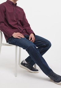 Camisa de cuadros roja y azul marino con botones, jeans de mezclilla azul y zapatillas oscuras con detalles blancos, sentado en una silla blanca.