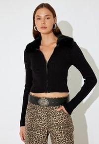 Schwarze gerippte, kurz geschnittene Strickjacke mit einem Kragen aus Kunstfell und silbernem Reißverschluss. Kombiniert mit beige-braunen Leopardenmusterhosen und einem schwarzen Gürtel.