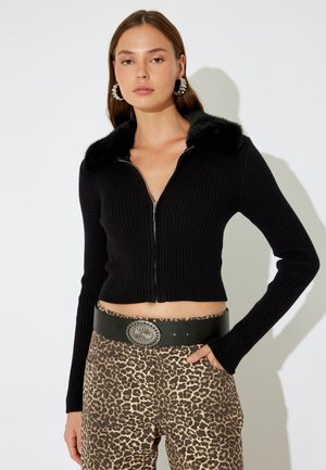 Schwarze gerippte, kurz geschnittene Strickjacke mit einem Kragen aus Kunstfell und silbernem Reißverschluss. Kombiniert mit beige-braunen Leopardenmusterhosen und einem schwarzen Gürtel.