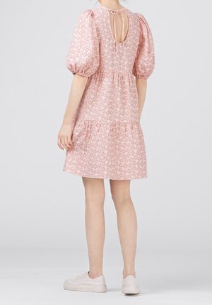 Kobieta ubrana w krótką różową sukienkę w kwiaty z bufkami na rękawach i wiązaniem z tyłu, w połączeniu z białymi trampkami, stojąca na jednolitym szarym tle.