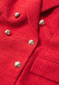Blazer texturé rouge vif avec boutons argentés brillants en relief et une poche frontale visible. Aucune personne présente.