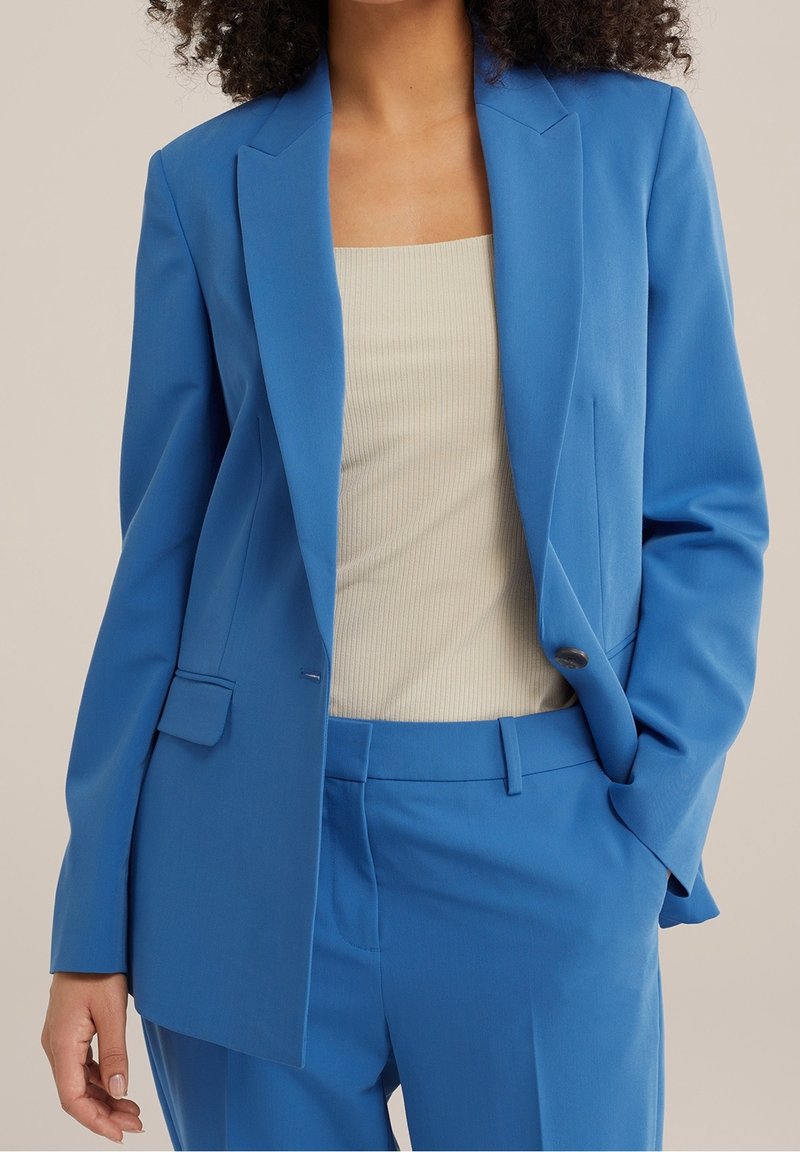 Femme portant un costume bleu sur mesure et un top sans manches côtelé beige, une main dans la poche, sur un fond neutre uni.