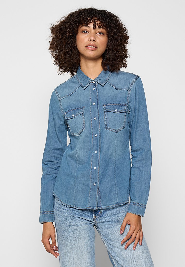 Chemise en denim bleu clair, dotée d'un col pointu, de deux poches poitrine et d'une fermeture par boutons. Texture lisse avec des détails de couture contrastés.