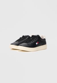 Fekete bőr sneaker, krém színű talppal, kerek fűzőkkel és egy logóval, amely piros, fehér és kék díszítéssel rendelkezik az oldalán.