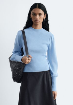 Femme aux longs cheveux foncés portant un pull bleu clair et une jupe noire, tenant un sac à épaule noir tressé sur un fond clair uni.