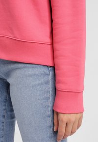 Sweatshirt rose avec des poignets côtelés, porté sur un jean bleu clair. Le tissu semble doux et texturé. Les bordures des poignets sont soigneusement finies.