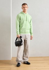Hoodie vert clair avec cordons de serrage, poche avant, associé à un jean large gris clair et des sneakers noires, tenant un petit sac noir.