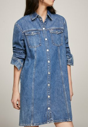 Frau trägt ein blaues Jeanskleid mit Knöpfen, hochgekrempelten Ärmeln und Brusttaschen, steht vor einem schlichten Hintergrund.