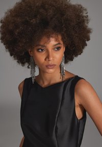 Frau mit voluminösen lockigen Haaren, die ein ärmelloses schwarzes Kleid und lange funkelnde silberne Ohrringe trägt, und leicht zur Seite blickt.