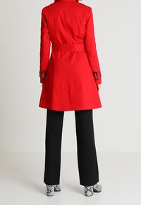 Červený trenčkot s páskem v pase, ramenními epaulety a dlouhými rukávy, kombinovaný s černými kalhotami s širokými nohavicemi a vzorovanými botami.