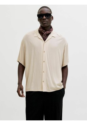 Mann trägt übergroßes beigefarbenes Hemd mit Knöpfen, schwarze Hose, schwarze Sonnenbrille und ein gemustertes braunes Halstuch vor schlichter Hintergrund.