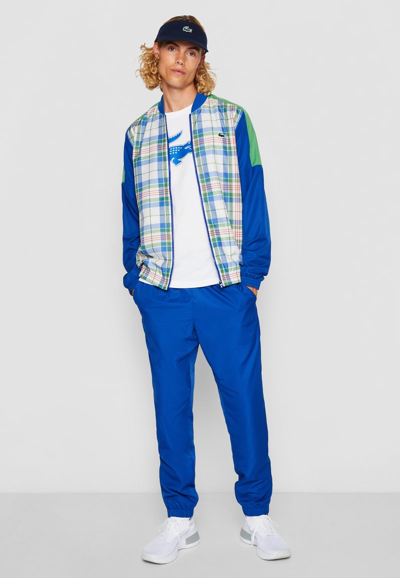 Survêtement bleu avec une veste bomber à carreaux aux manches vertes, t-shirt blanc avec logo bleu, et baskets blanches.