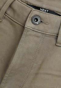 Pantaloni din bumbac bej, cu o textură netedă, având un buton metalic negru și cusături pentru durabilitate. Eticheta cu logo este vizibilă lângă talie.