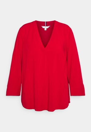 Blouse rouge à col en V avec des manches longues, fabriquée dans un tissu lisse. possède une coupe ample, des fentes sur les côtés et un ourlet droit. Étiquette visible au niveau du col.