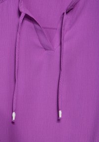 Lilla pullover lavet af tekstureret stof, med en v-hals og to hvide snore fastgjort ved halsudskæringen.
