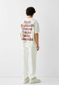 Camiseta blanca de gran tamaño con texto marrón que enumera nombres de ciudades en la parte trasera, combinada con pantalones blancos a juego y zapatillas beige.