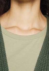 Olive green knit cardigan layered over a light green cotton t-shirt, featuring a round neckline and visible stitching details.