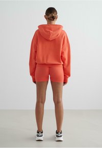 Koraalkleurige hoodie met capuchon, oversized pasvorm, gecombineerd met bijpassende fitted shorts. Witte sneakers maken de sportieve uitstraling compleet.