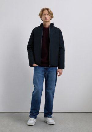 Jong persoon die binnen staat, gekleed in een zwarte jas, een donkere sweater, blauwe jeans en witte sneakers tegen een effen lichte achtergrond.