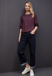 Blusa burdeos de manga larga con ribete blanco, jeans oscuros de corte relajado con dobladillos remangados y zapatillas blancas sobre un fondo gris.