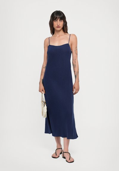 Vestido midi azul marino con tirantes finos, tejido suave y dobladillo acampanado. Modelo sosteniendo un bolso blanco y usando sandalias negras de dedo.