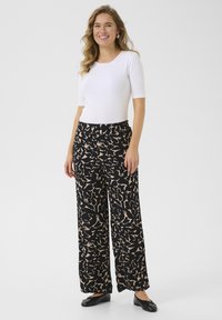 Un pantalon large en tissu noir avec un motif abstrait beige, associé à un haut ajusté à manches courtes blanc et des chaussures plates noires.