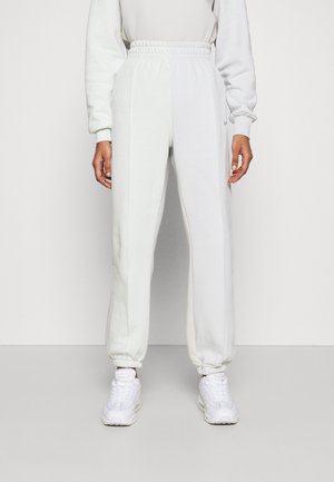 Osoba oblečená v bílých teplácích s elastickými manžetami, v kombinaci s bílými teniskami a bílým dlouhým rukávem, stojící na jednobarevném pozadí.