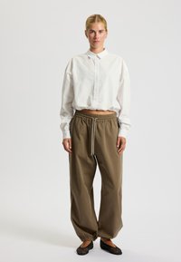 Camicia bianca con colletto e maniche lunghe, dalla vestibilità ampia. Pantaloni marroni con vita elasticizzata e cordoncino. Scarpe nere slip-on con design in rete.
