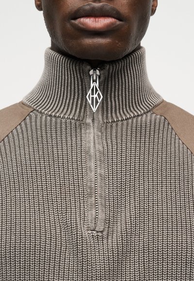 Κοντινό πλάνο ενός ατόμου που φοράει γκρι πλεκτό πουλόβερ με ψηλό ριμπ κολάρο και ασημί φερμουάρ σε σχήμα γεωμετρικού διαμαντιού.
