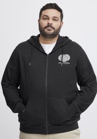 Svart zip-up hettegenser laget av bomullsblanding, med frontlomme, ribbede mansjetter og et grafisk trykk på venstre bryst.
