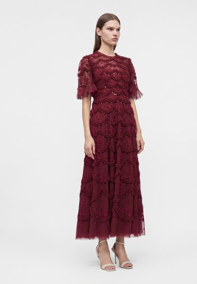 Femme portant une robe longue en dentelle bordeaux avec des couches festonnées et des manches courtes transparentes, associée à des sandales beiges à talons hauts.