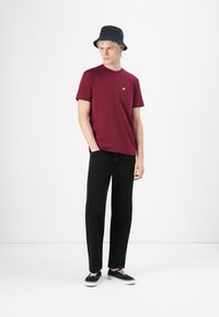 Maronfarvet bomulds T-shirt med brystlomme, parret med sorte lige ben bukser og sorte sneakers. Modellen bærer en sort bøttehat.