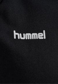 Tejido negro con una trama simple y un destacado logo blanco de "hummel" centrado en la tela. No hay patrones ni acentos adicionales visibles.