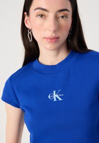 Une jeune femme aux longs cheveux noirs porte un t-shirt Calvin Klein bleu vif et de grandes boucles d'oreilles en argent, regardant directement l'objectif.