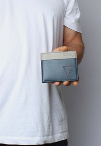 Blue leather wallet with a grey top edge, featuring a triangular cutout and red stitching detail, held in a hand against a plain background.