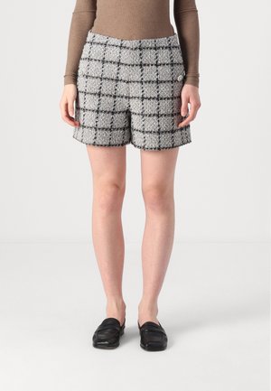 Femme portant un short en tweed à carreaux gris et noirs, un haut marron à manches longues et des mocassins noirs, debout devant un fond uni.