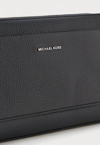 Sort tekstureret læderpung med sølv "Michael Kors" logo og lynlåslukning på en hvid baggrund.