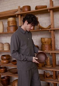 Grijze ritsjacket over een bijpassende outfit, met een gestructureerd oppervlak. Een persoon houdt een donkerhouten kom vast, omringd door houten artefacten op een plank.