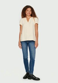 Top bege de mangas curtas com decote em V e uma textura subtil, combinado com jeans azuis ajustados e sapatos pretos robustos, de pé contra um fundo claro.
