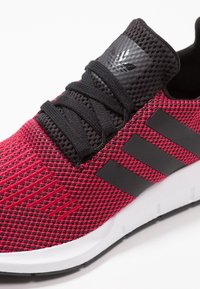 Röd och svart sportskor med en texturerad stickad ovandel, svarta snören och tre svarta ränder. Har en vit mellansula och gummisula.