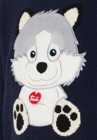 Indumento blu con un personaggio di lupo ricamato in peluche, con pelo grigio, faccia bianca e zampe nere, contrassegnato da un'etichetta rossa "Trudi".