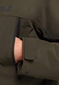 Giacca verde oliva con tasca con zip, tessuto a motivo e polsini regolabili. Logo visibile nell'area della spalla, il materiale è testurizzato.