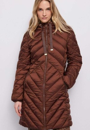 Donna che indossa un lungo cappotto imbottito marrone trapuntato con cappuccio e cerniera frontale, in piedi davanti a uno sfondo chiaro e uniforme.