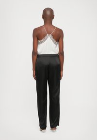 Persona stāv, ģērbta baltā bezpiedurņu topā ar mežģīņu apdari un melnās satīna platās biksēs, ar bēšiem papēžiem uz neitrāla fona.