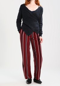 Pull en maille gris foncé avec de longues manches et un col en V, associé à un pantalon large à rayures bordeaux et des chaussures plates noires.