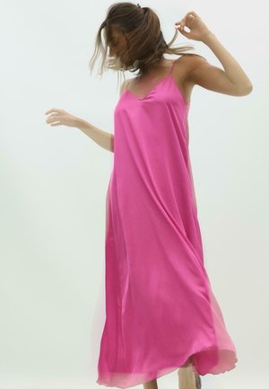 Femme aux cheveux bruns mi-longs portant une robe fluide rose vif sans manches sur un fond clair uni.