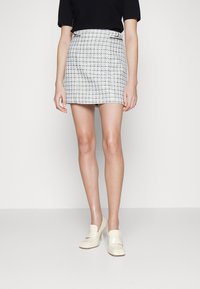 Mujer vestida con una falda corta a cuadros blancos y azules, top negro y zapatos de tacón crema, de pie contra un fondo blanco liso.