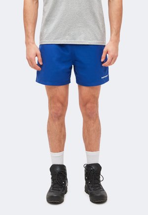 Άνδρας που φορά μπλε σορτς, γκρι μπλουζάκι, λευκές κάλτσες και μαύρα ορειβατικά παπούτσια, στέκεται μπροστά σε απλό φωτεινό φόντο.