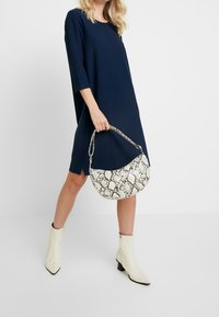Granatowa, teksturowana sukienka o kroju shift z rękawami do połowy łokcia; trzyma białą torebkę z motywem skóry węża; kremowe botki za kostkę.