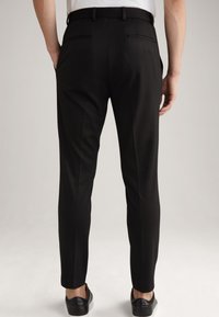 Schwarze maßgeschneiderte Hose mit schlanker Passform, zwei Gesäßtaschen und einer glatten Stoffstruktur. Kombiniert mit schwarzen Slippers.